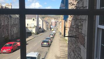 Vista de la calle a través de una ventana, con edificios de piedra, coches aparcados y cielo despejado.
