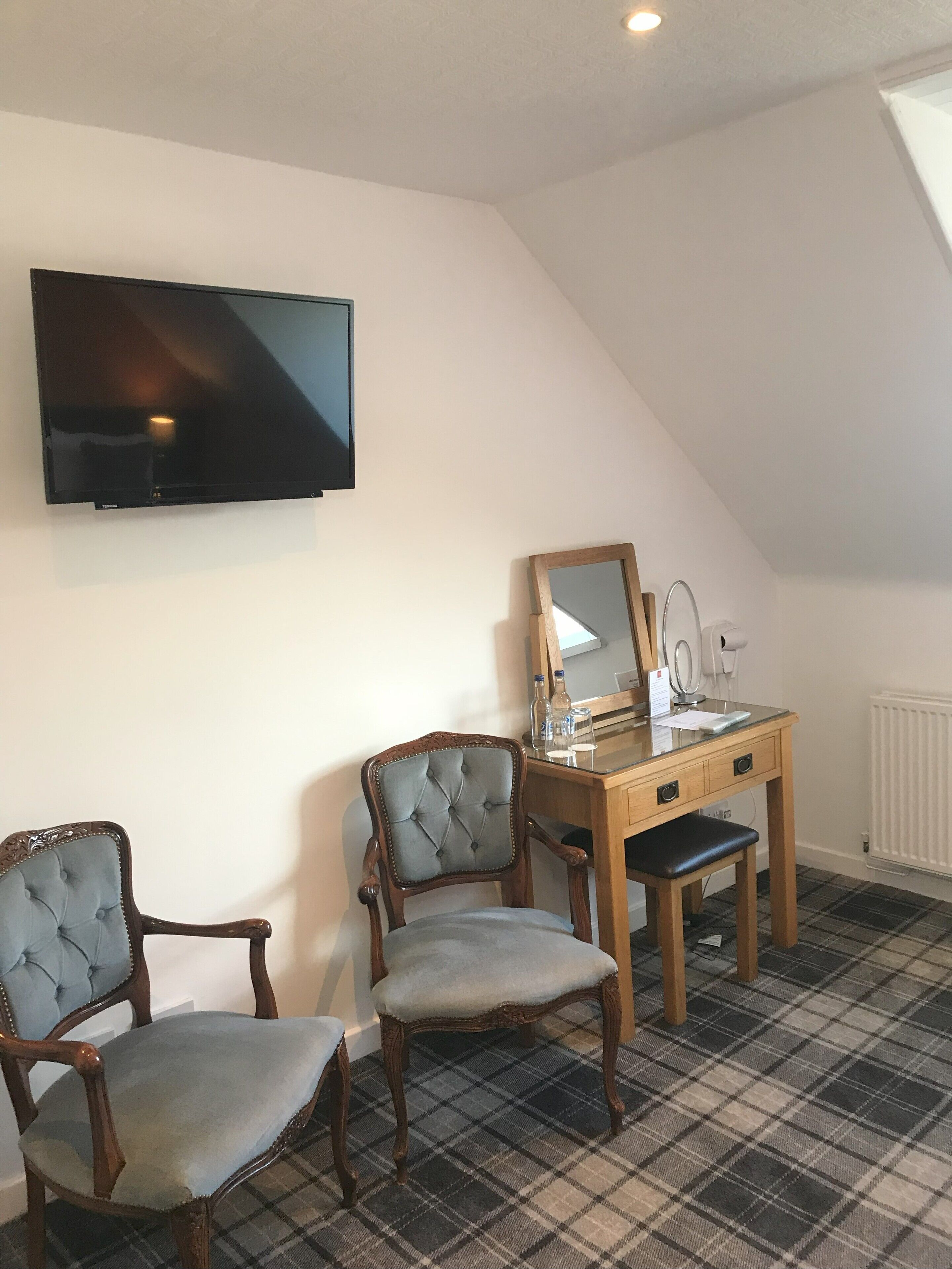 Una habitación con televisión montada en la pared, un escritorio con espejo, dos sillones tapizados y una alfombra a cuadros.