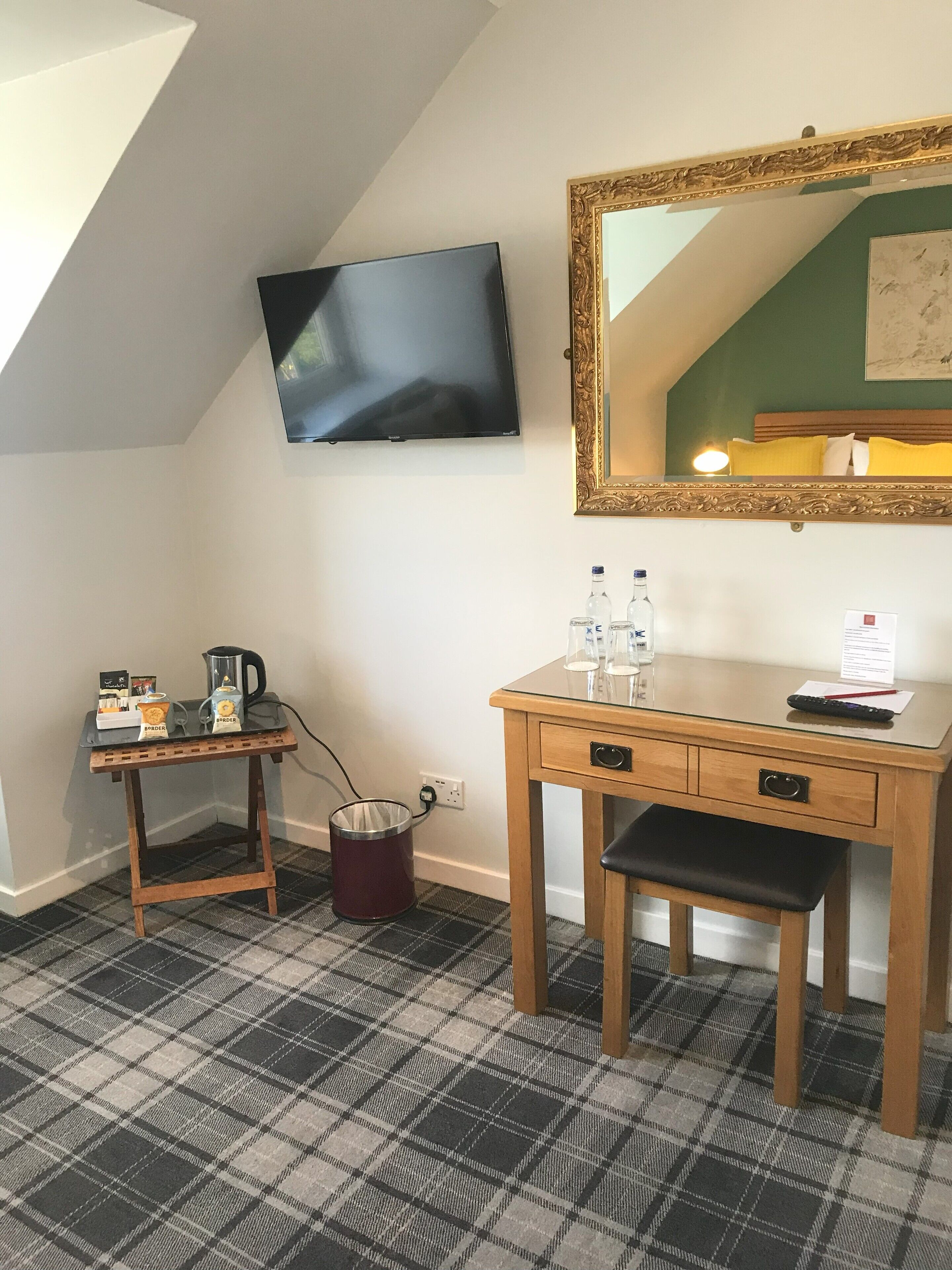 A hotel room with a wooden desk, a chair, a television mounted on the wall, a mirror with an ornate frame, and a plaid-patterned carpet.