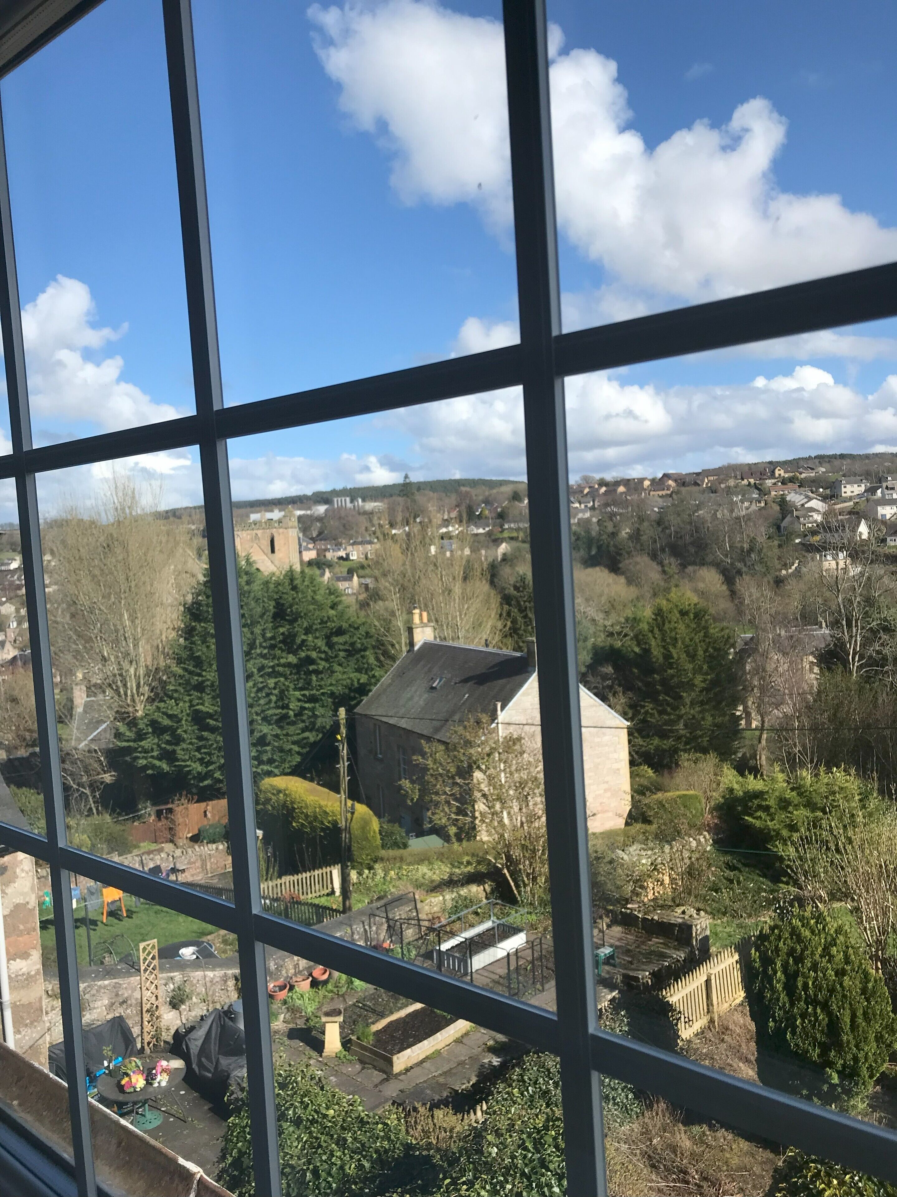 A view through a window with a grid pattern, overlooking a residential area with houses, trees, and a distant hill.