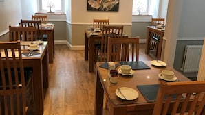 A dining area with wooden tables and chairs, a painting on the wall, and windows with views of greenery. - Meadhon House (Jedburgh)