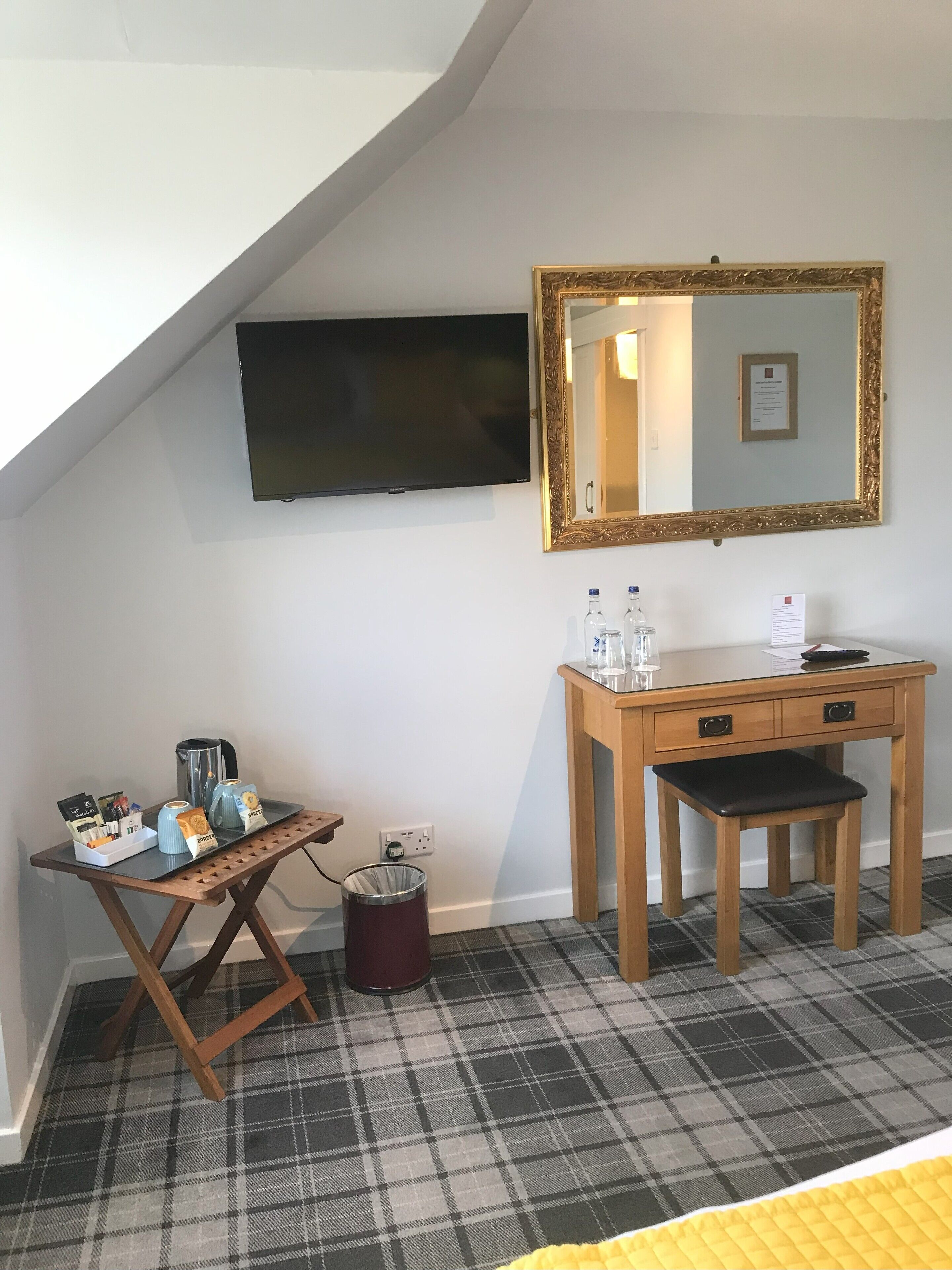 A hotel room with a flat-screen TV mounted on the wall, a wooden desk with a chair, a small table with a tray of breakfast items, and a trash can.