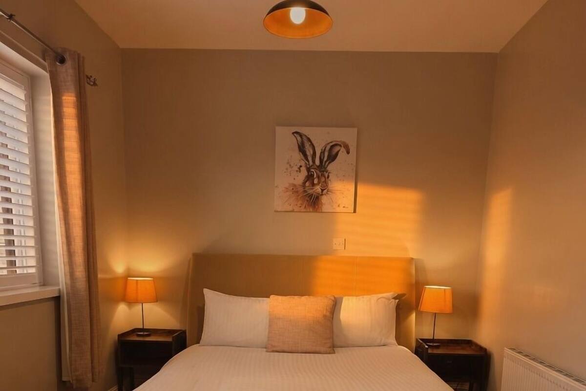 Un dormitorio con una cama grande, dos mesitas de noche con lámparas, un cabecero, una ventana con persianas y una obra de arte colgada en la pared.