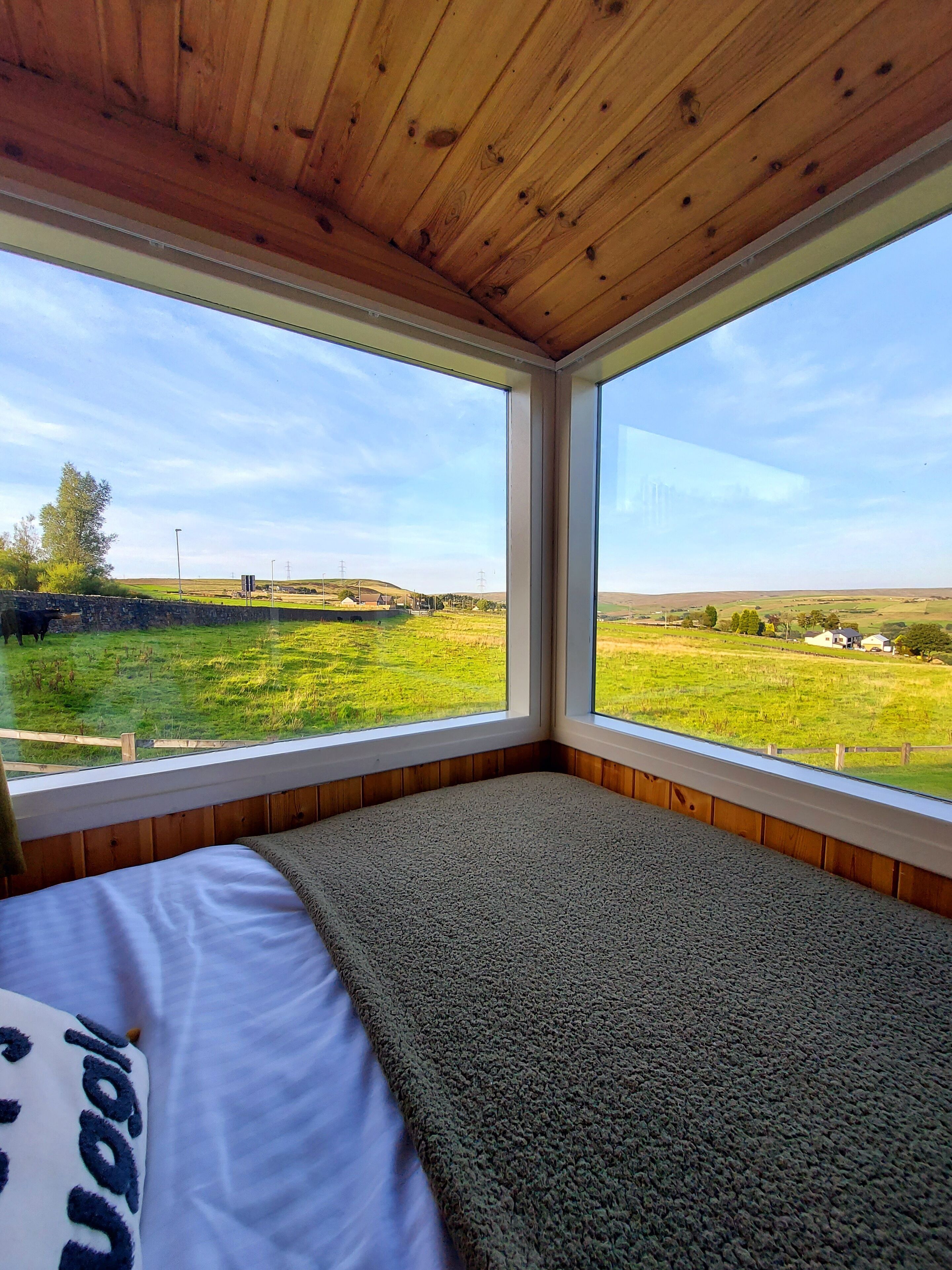 A bedroom with a large window, wooden ceiling, and a view of a green field.