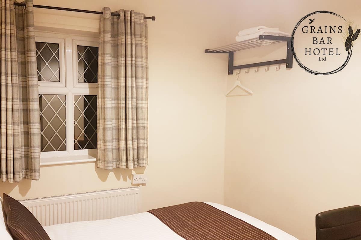 Un dormitorio con cama, ventana con cortinas, radiador, estante en la pared con toallas y un letrero para Grains Bar Bar Hotel Ltd.