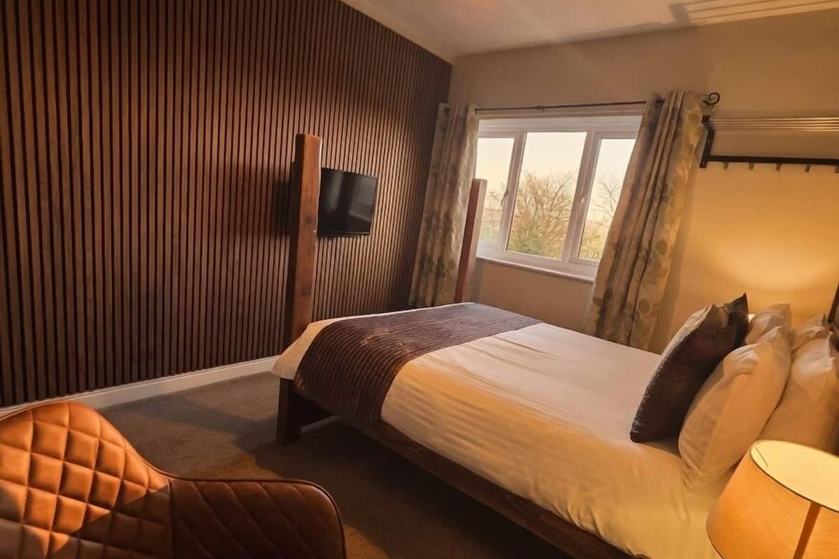 Una habitación de hotel con una cama, un sillón de cuero, una mesita de noche con una lámpara, un televisor montado en la pared y una ventana con cortinas.