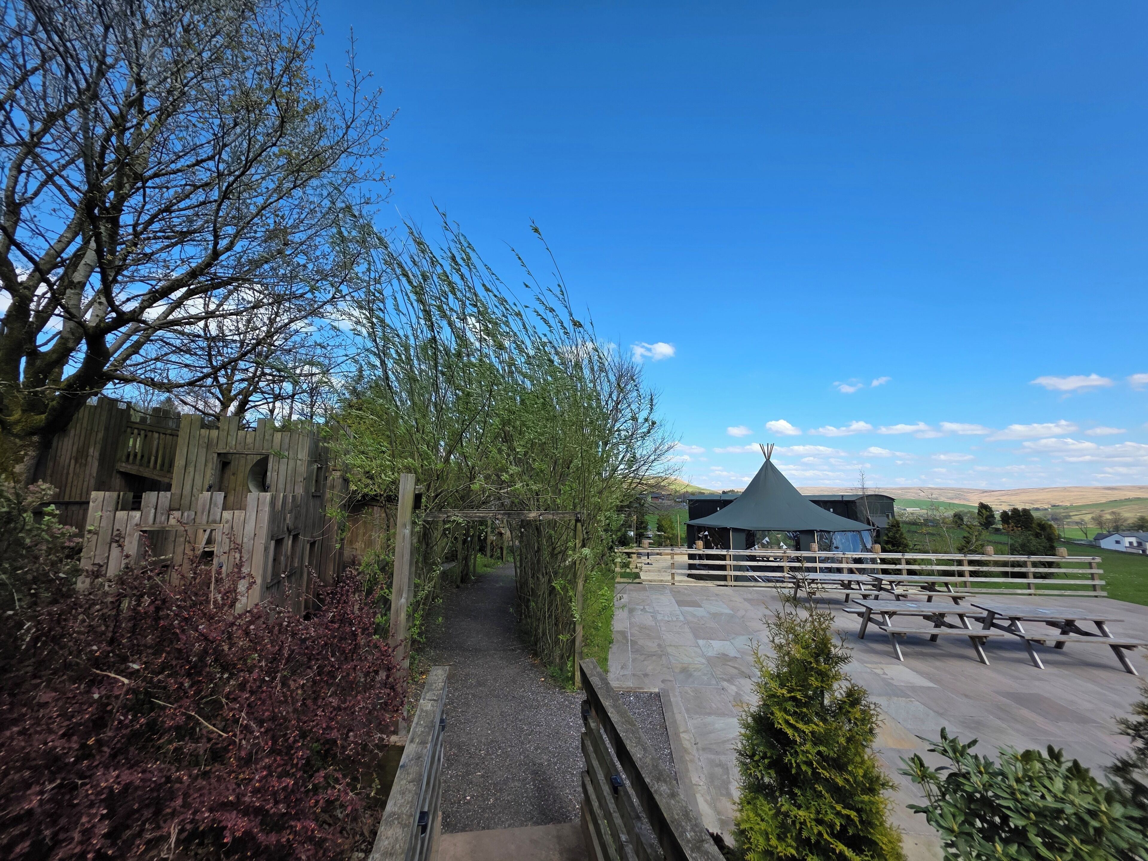 A pathway leading to a covered seating area with picnic tables, surrounded by wooden fences and vegetation.