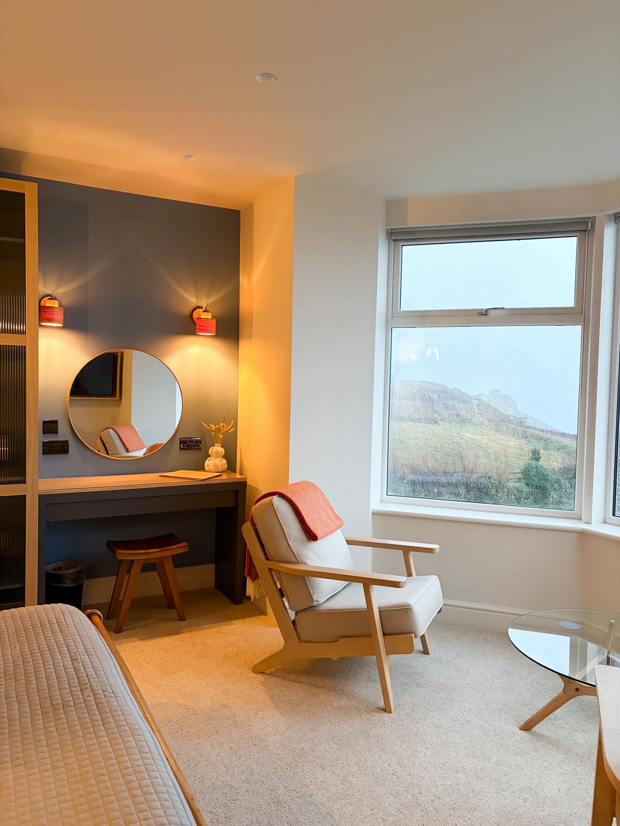 A bedroom with a bed, a desk with a chair, a round mirror, and a window with a view of a hill.
