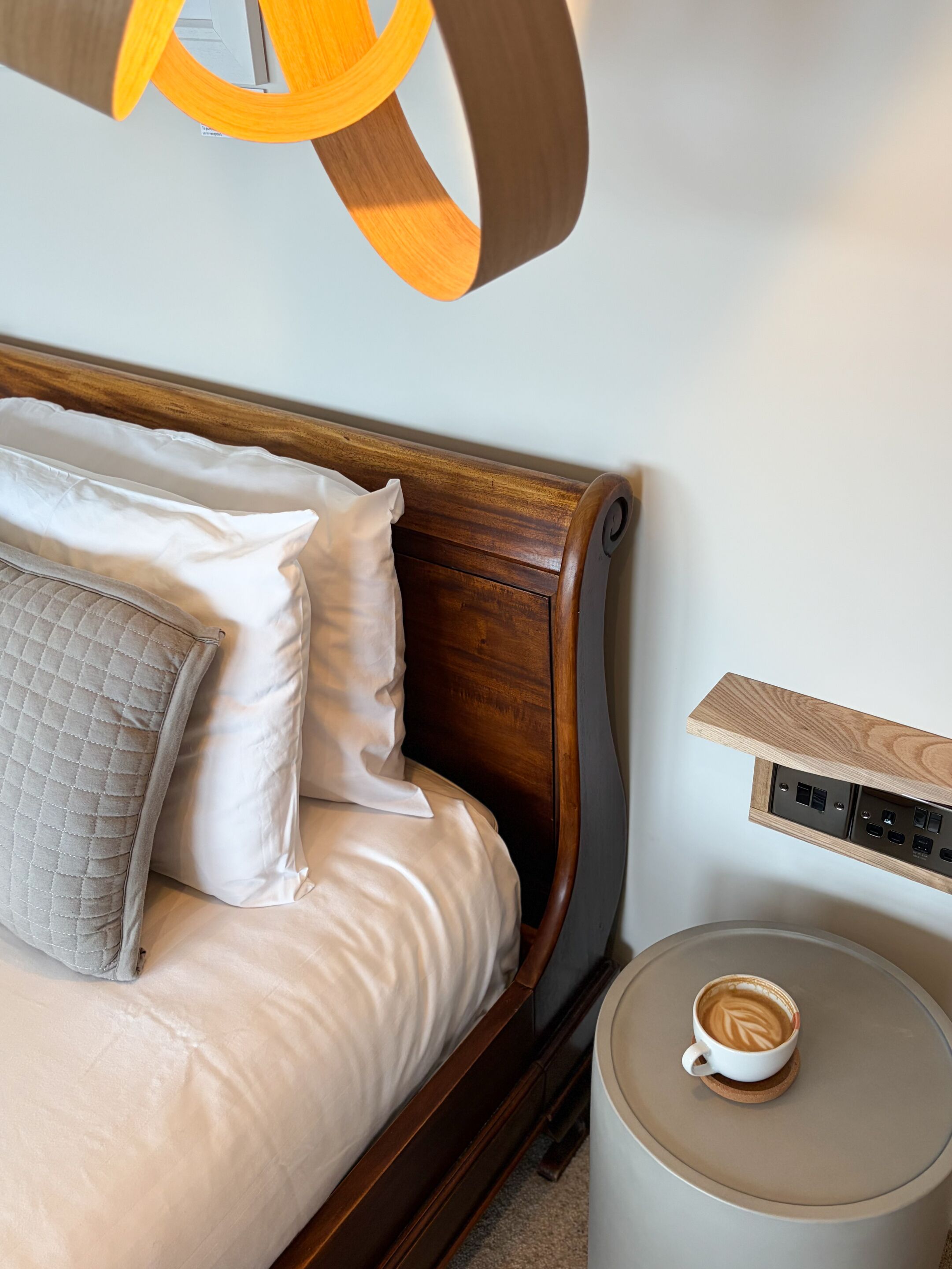 Una cama con cabecera de madera, un almohadón acolchado y una almohada blanca. Se ve una mesita de noche con una taza de café y una regleta de enchufes.