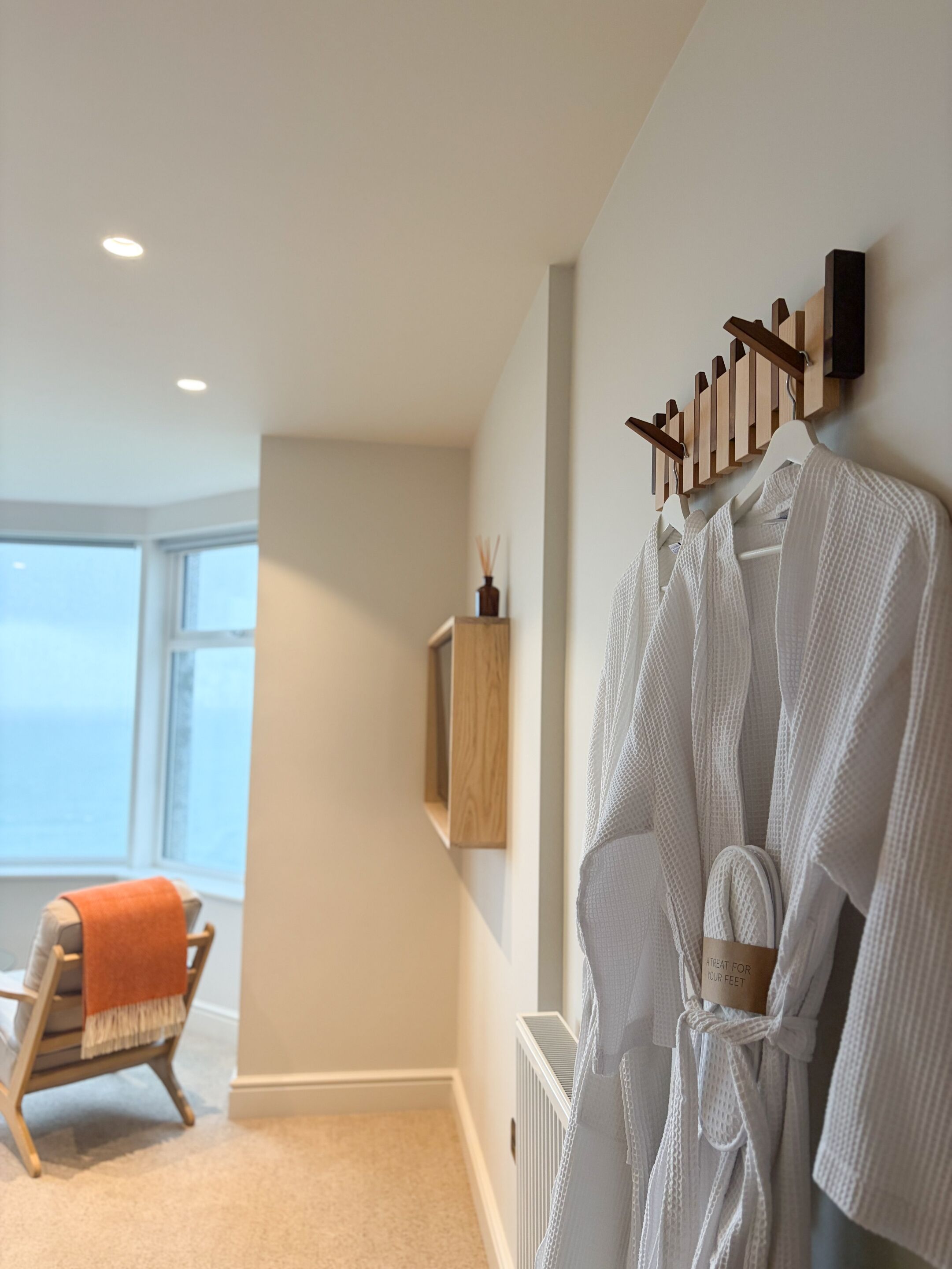 A hotel room with a large window, a wooden chair with an orange throw, and a white robe hanging on a rack.