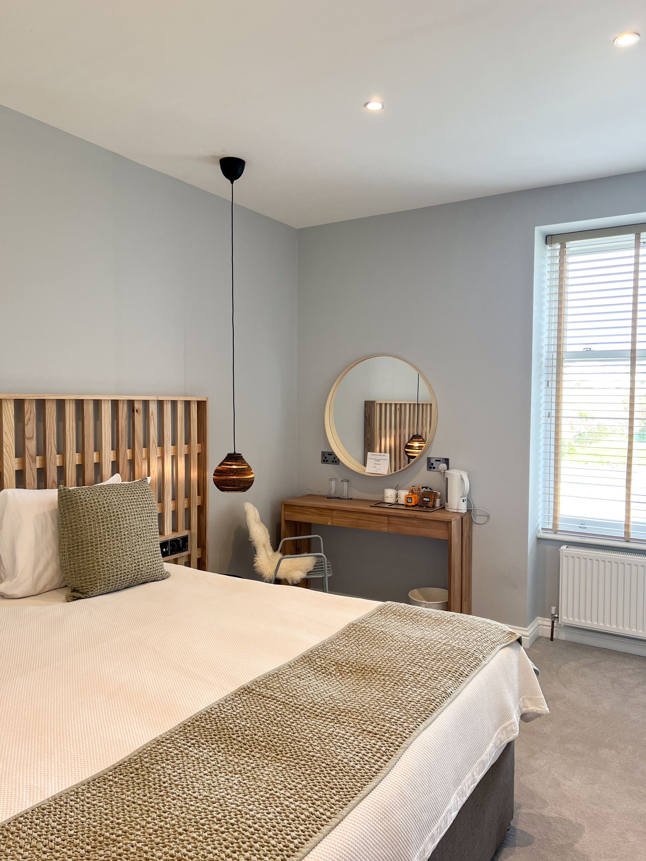 Un dormitorio moderno con cabecera de madera, un espejo redondo, un escritorio con silla y un ventanal con persianas.
