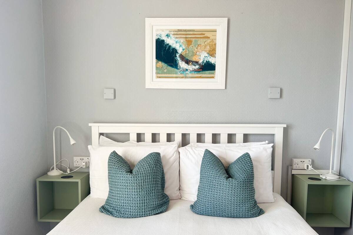 A neatly made bed with white and teal pillows, a framed map on the wall, and bedside tables with lamps and charging ports.
