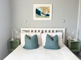 Una cama bien hecha con almohadas blancas y azul turquesa, un mapa enmarcado en la pared y mesitas de noche con lámparas y puertos de carga.
