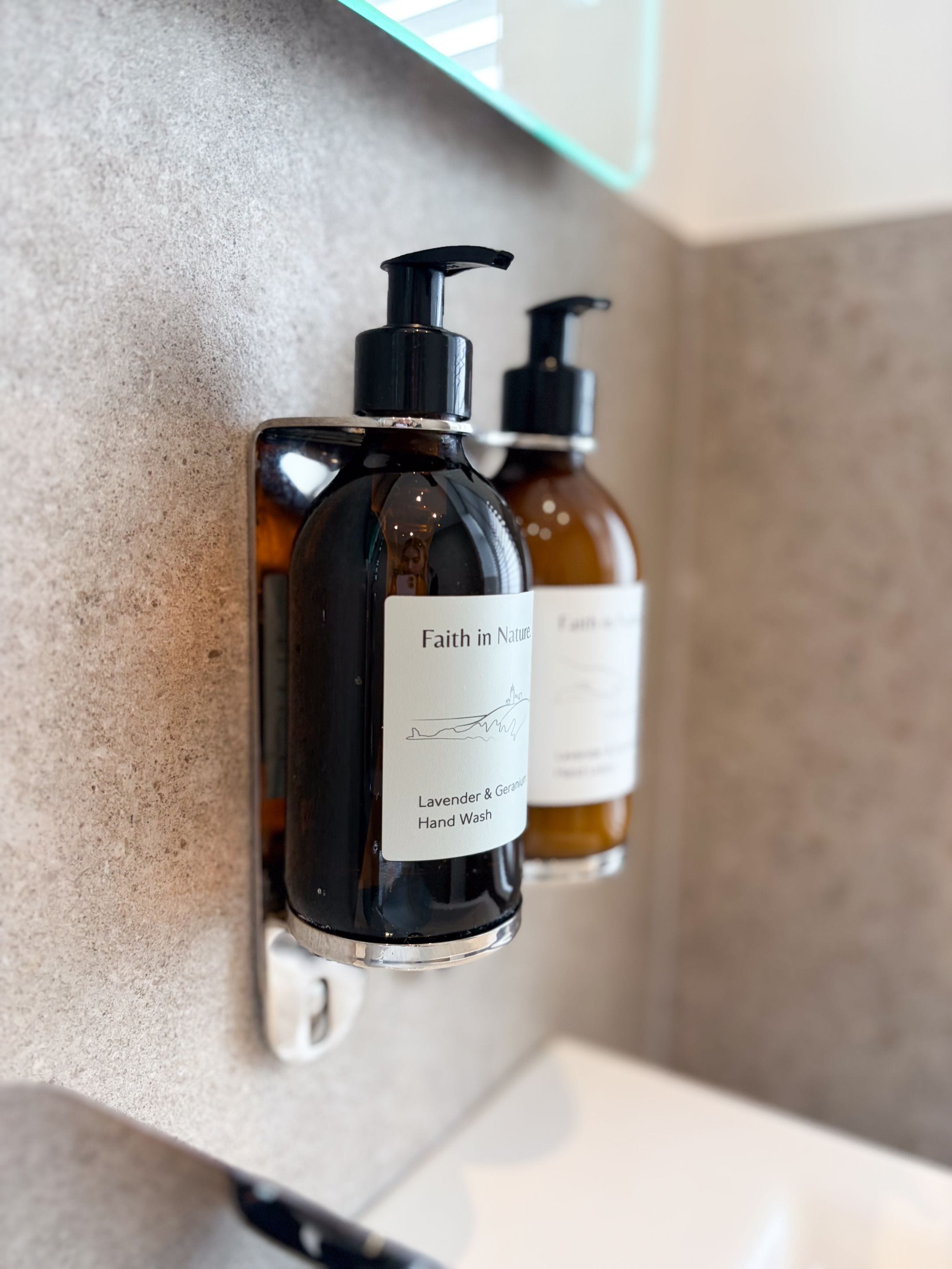 Two hand wash bottles mounted on a wall, one labeled 'Faith in Nature' with lavender and geranium scents.