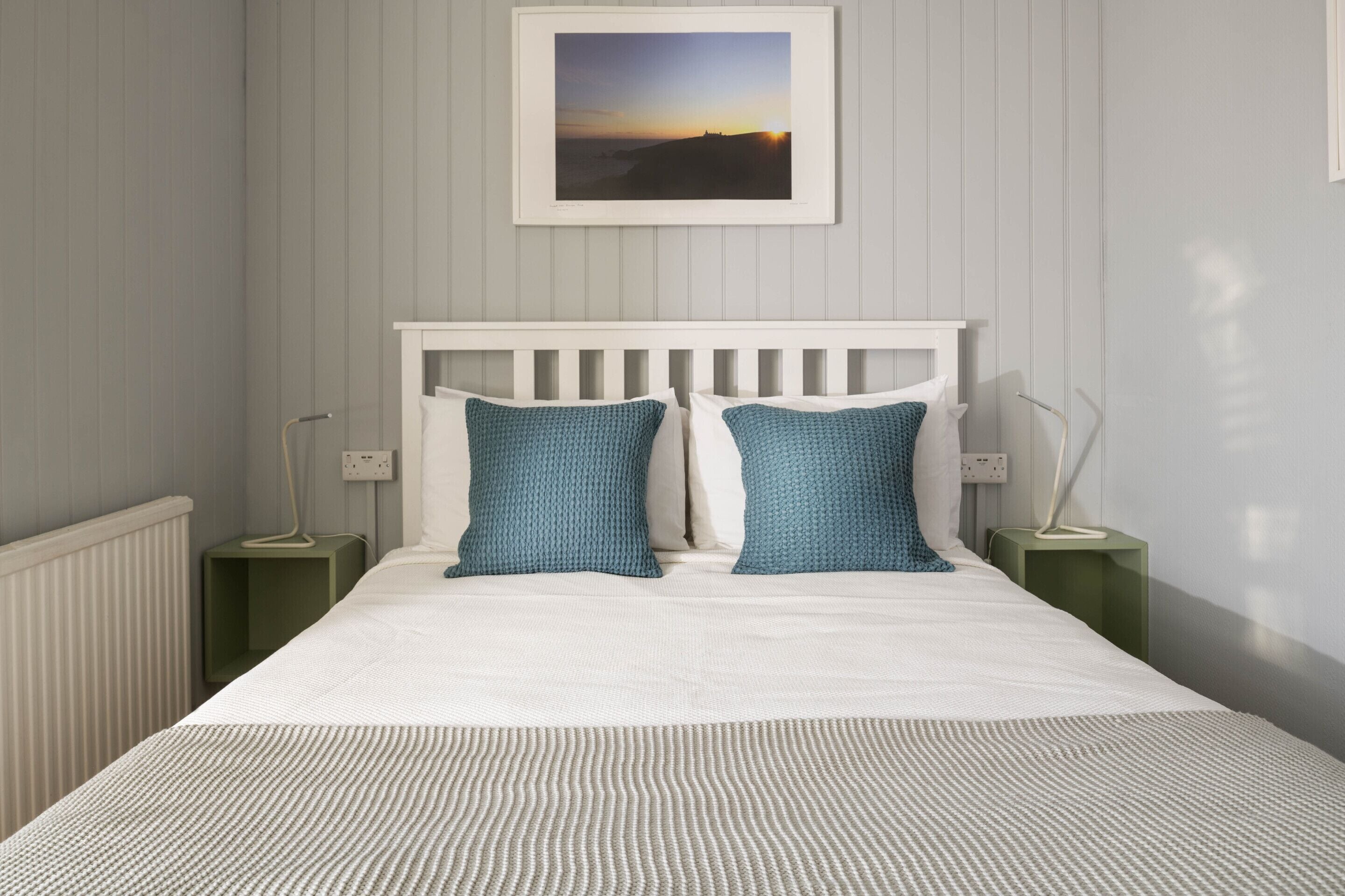 A bedroom with a white bed, two blue pillows, a framed picture on the wall, and a radiator.