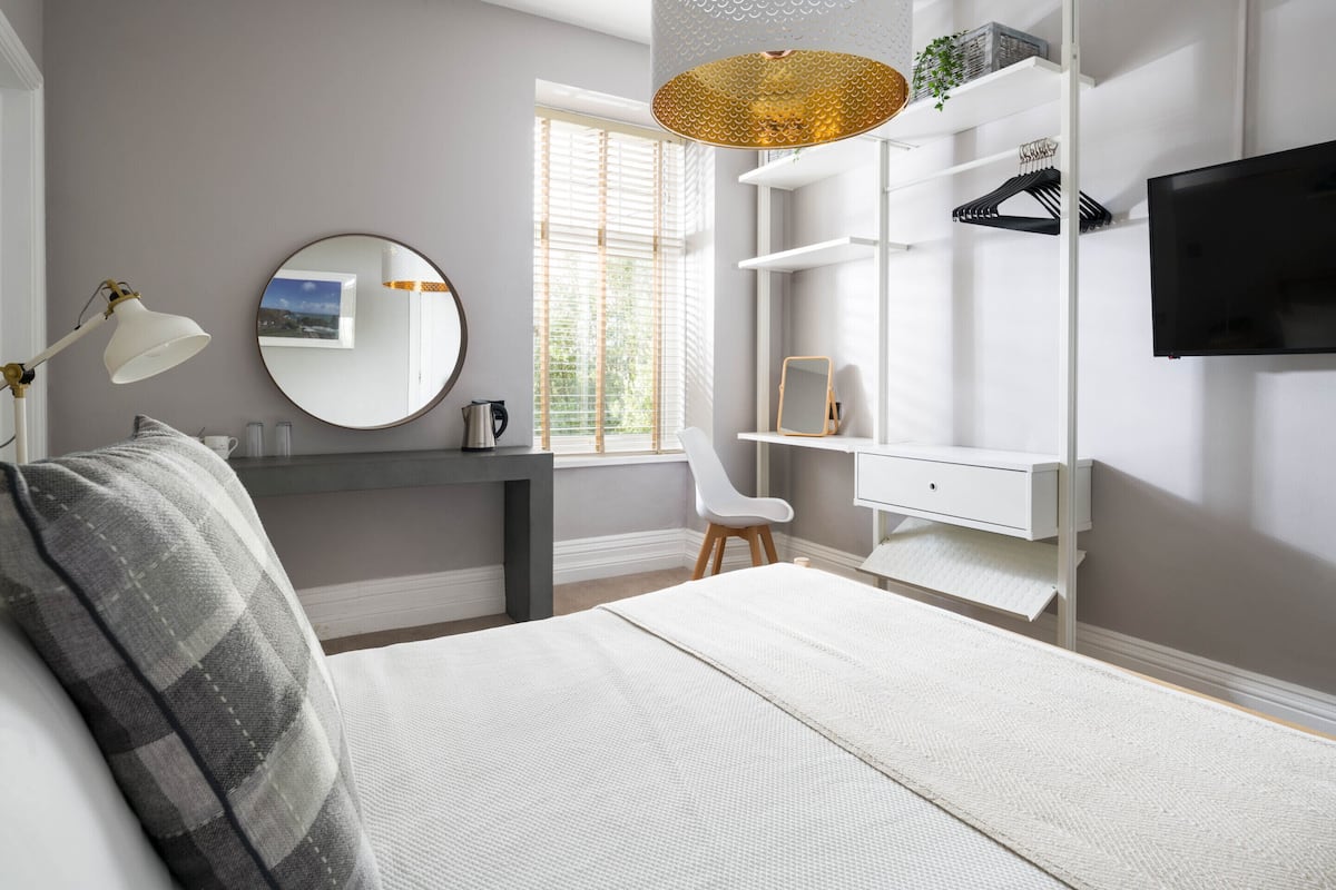 A bedroom with a bed, a desk, a chair, a TV, and a window with blinds.