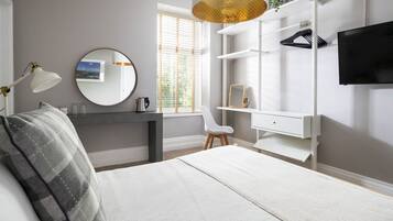 A bedroom with a bed, a desk, a chair, a TV, and a window with blinds.