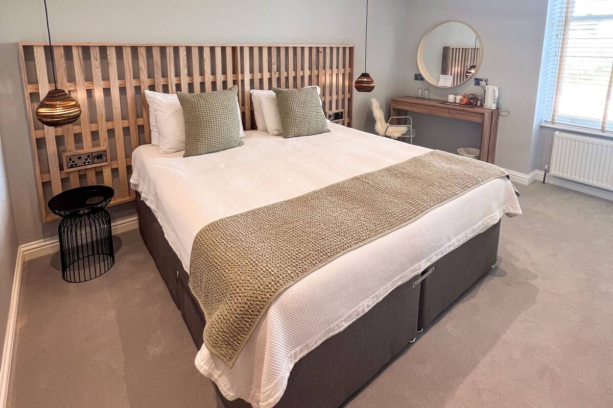 A modern bedroom with a large bed, a wooden headboard, a bedside table, a mirror, and a window with blinds.