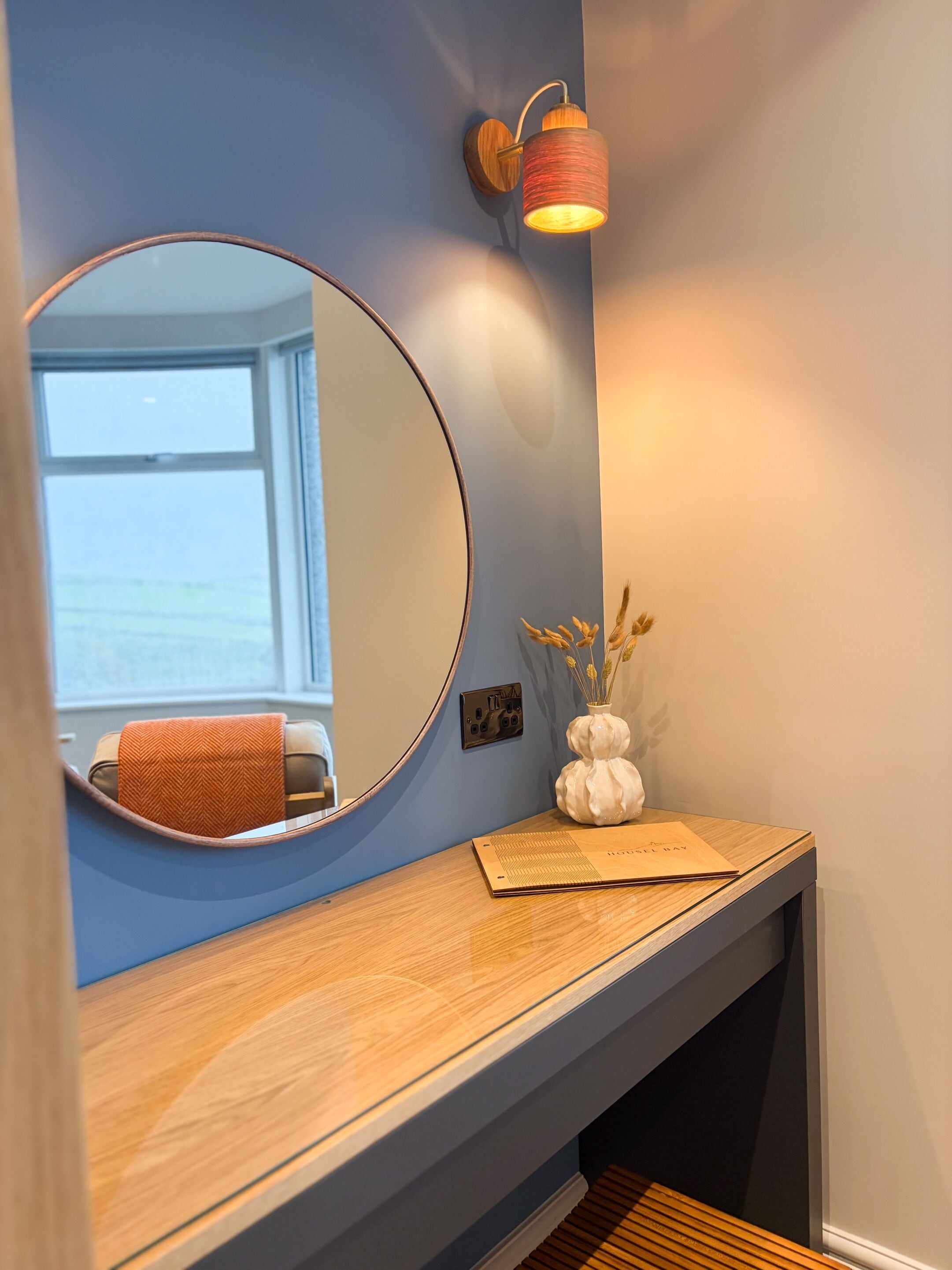 A bathroom with a round mirror, a wooden vanity, a wall-mounted light fixture, and a window with a view of the sea.