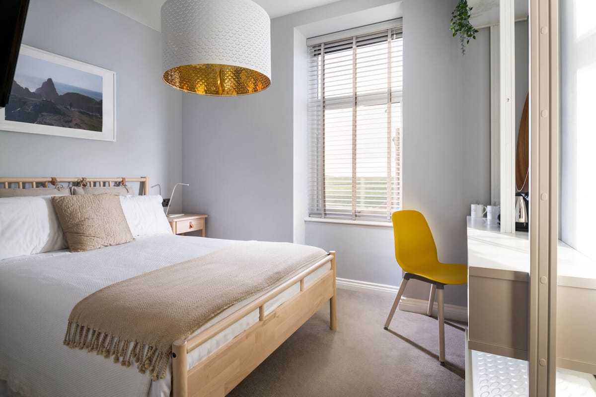 A bedroom with a bed, bedside table, lamp, window with blinds, and a chair.