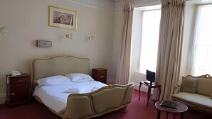 Egy szállodai szoba, amelyben egy nagy ágy, egy éjjeliszekrény telefonnal, egy kis asztal laptopmal, egy kanapé és egy függönnyel ellátott ablak található.