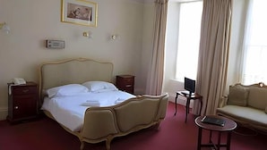 A hotel room with a large bed, a bedside table with a phone, a small table with a laptop, a sofa, and a window with curtains.