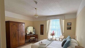 A bedroom with a bed, a desk with a mirror, a wardrobe, a chandelier, and a window with curtains.