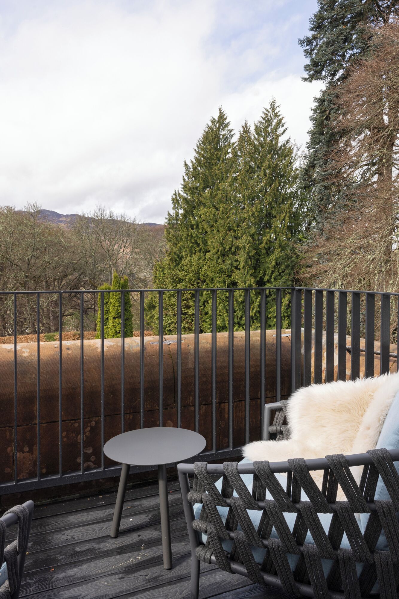 A balcony with a metal railing, a round table, and a cushioned chair with a fur throw.