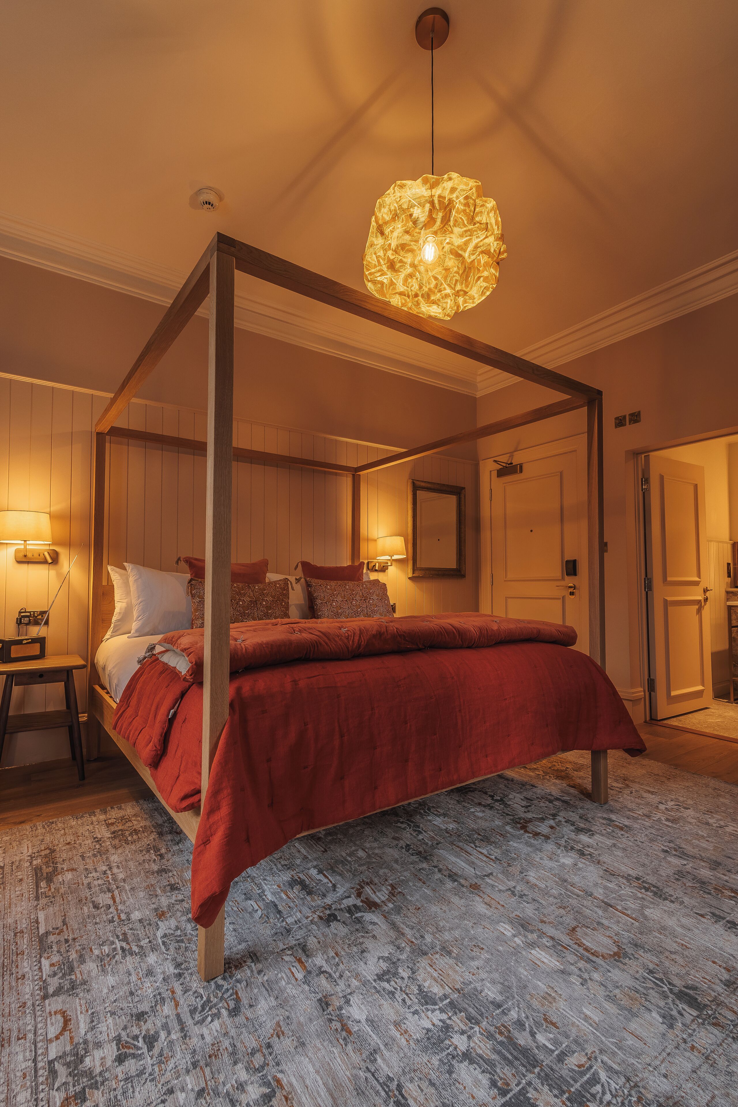 A four-poster bed with a red bedspread, a wooden floor, and a ceiling light fixture.