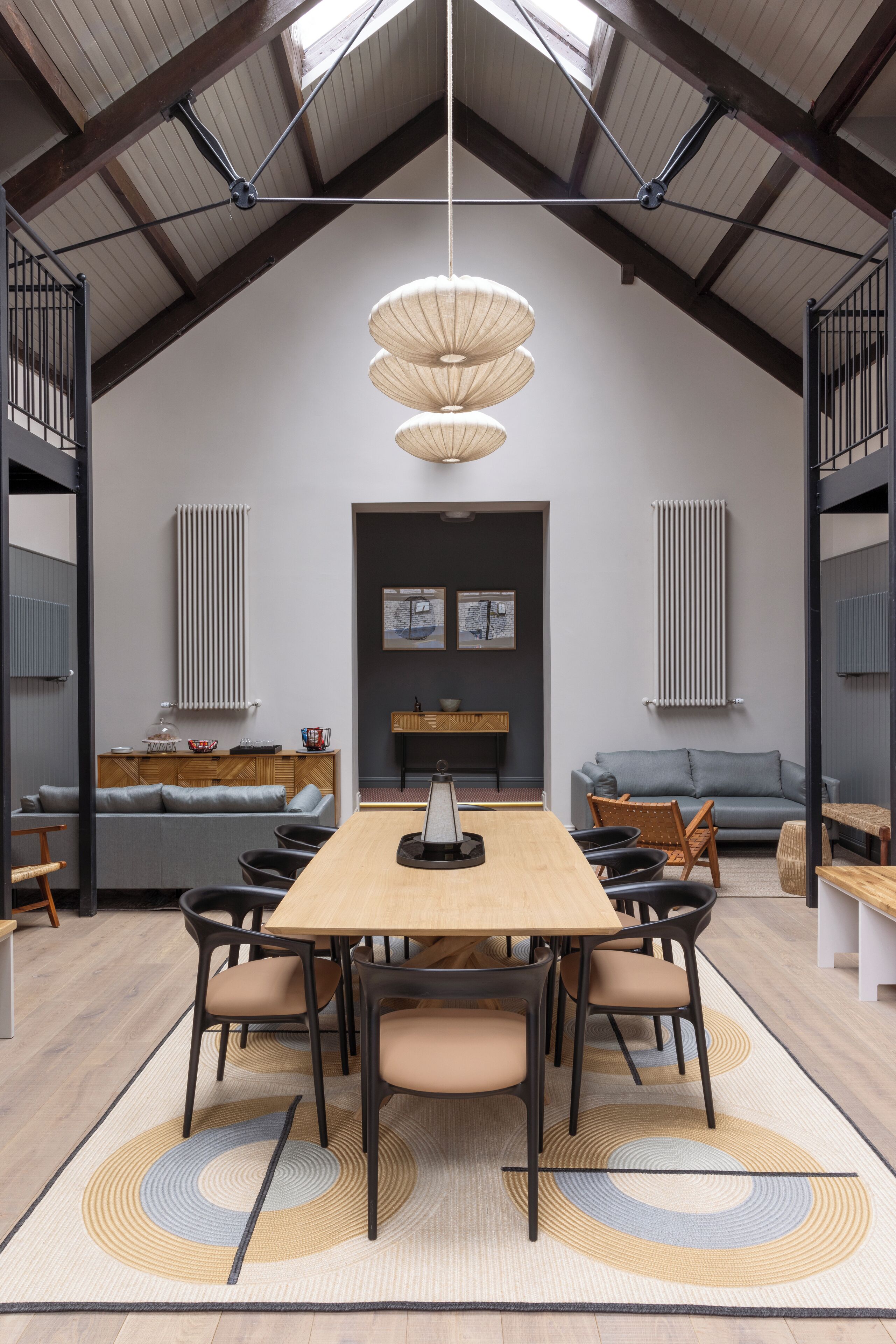 A modern dining area with a wooden table and chairs, a central light fixture, and a view of the kitchen and living area.
