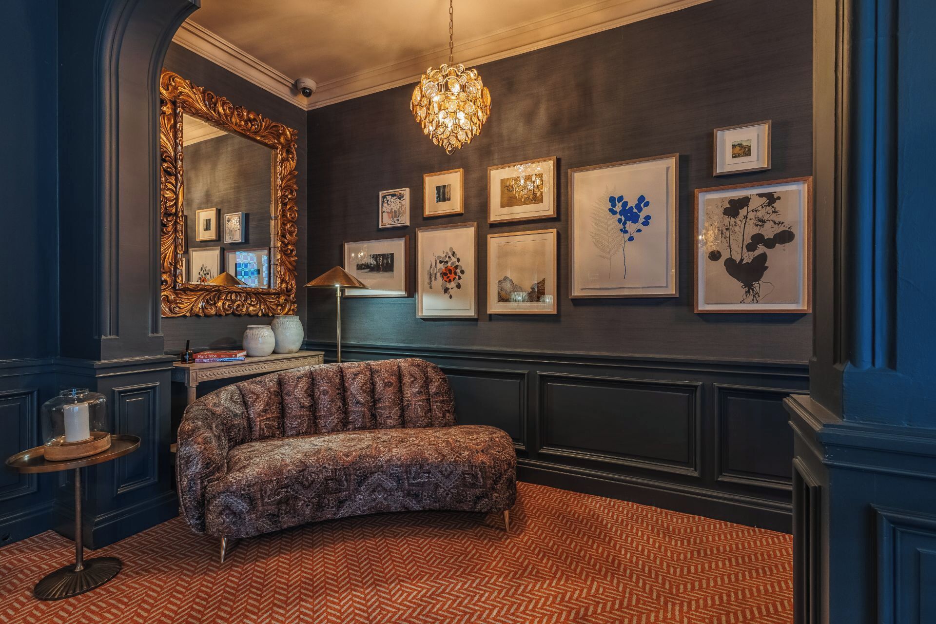 Una habitación con una pared oscura, un espejo grande y ornamentado, un sofá estampado y un suelo con estampado de espiga.