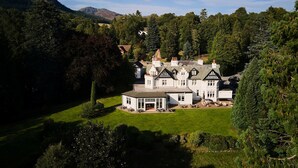 Un edificio grande de estilo Tudor, blanco y con múltiples chimeneas, rodeado de exuberante vegetación y montañas al fondo.