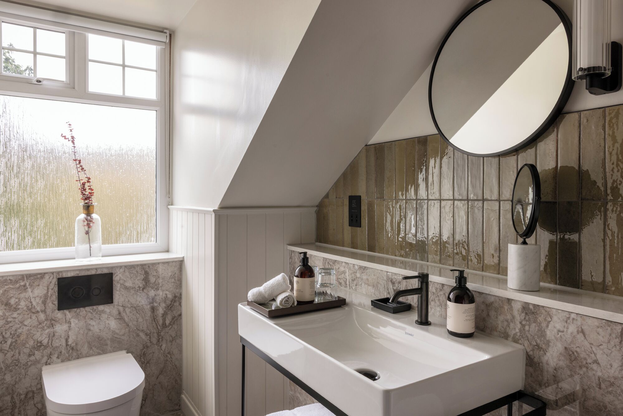 Une salle de bain avec un lavabo blanc, un miroir et une fenêtre agrémentée d’un vase de fleurs.