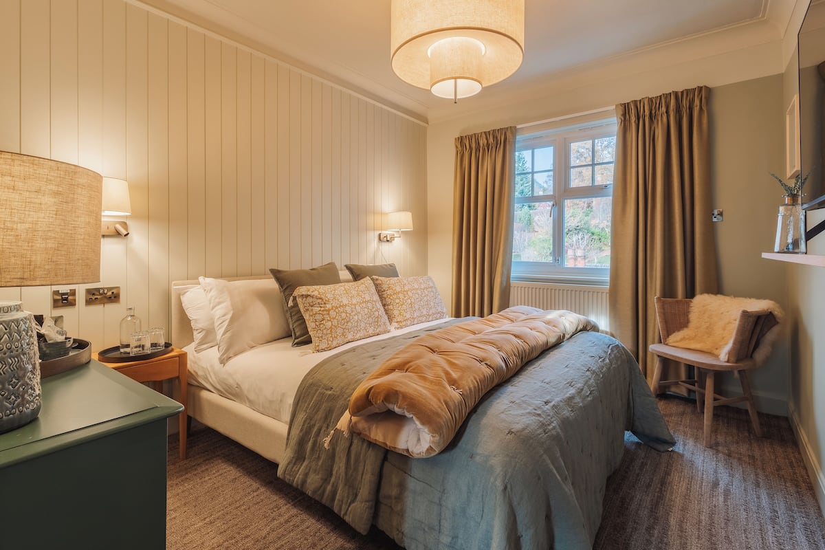 A bedroom with a large bed, bedside lamps, a window with curtains, a chair, and a small table with a vase.