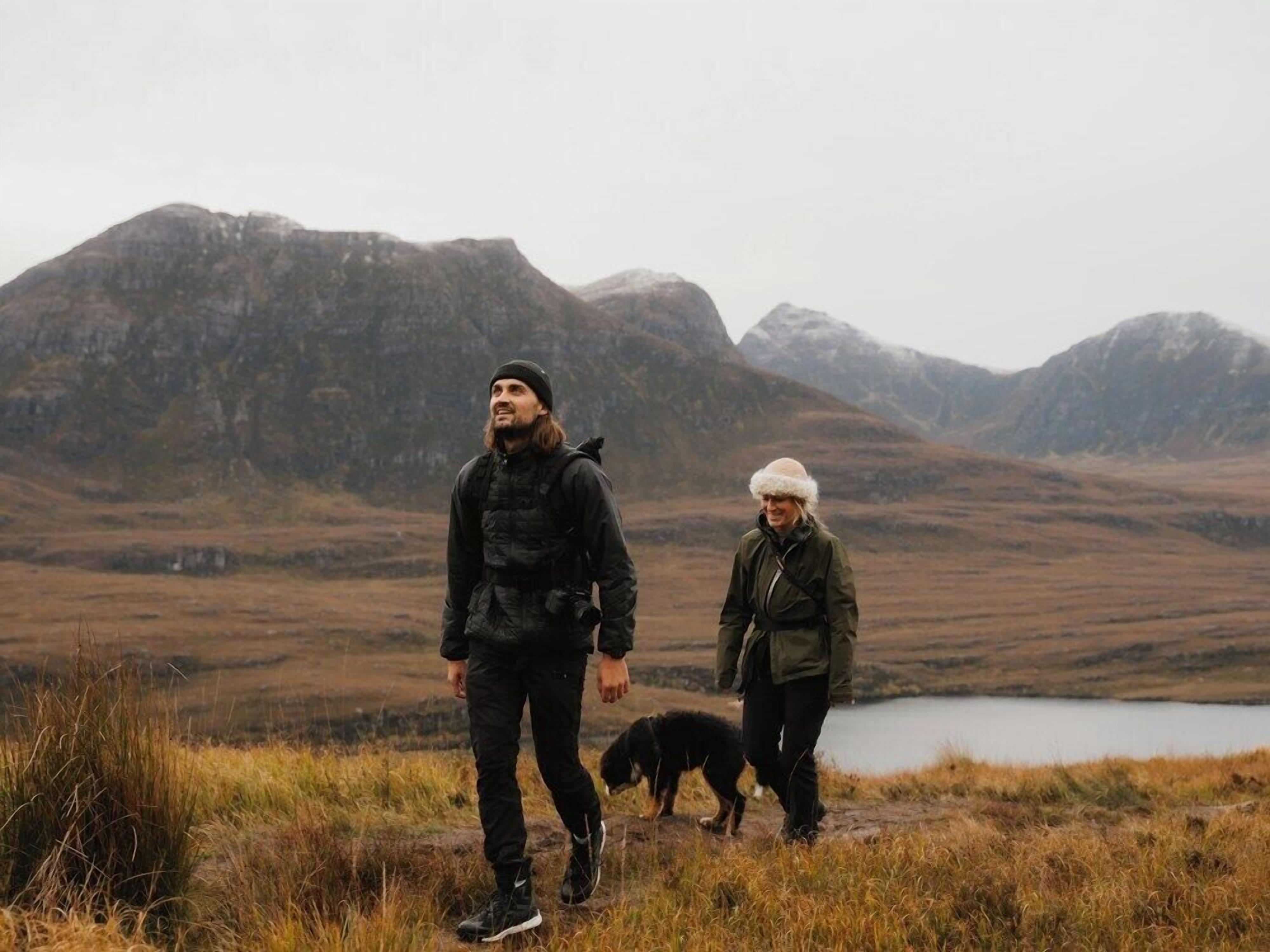 Two people walking a dog in a mountainous area with a lake in the background.