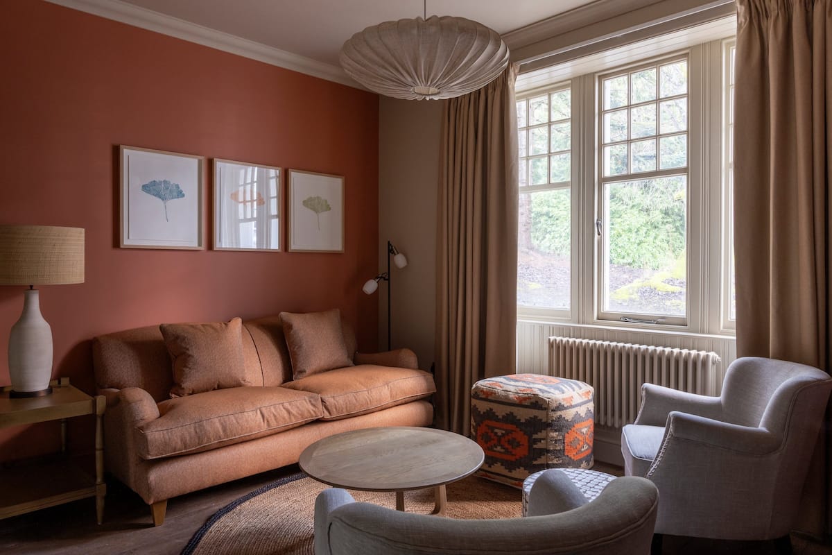 A cozy living room with a sofa, armchairs, a round coffee table, and a patterned ottoman. There are framed pictures on the wall, a lamp on a side table, and a window with curtains.
