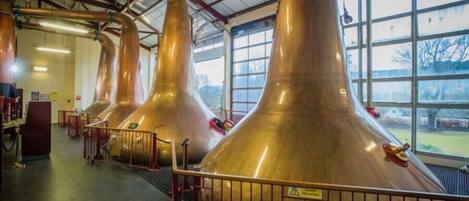 A distillery interior with large copper stills, a metal railing, and a view through large windows.