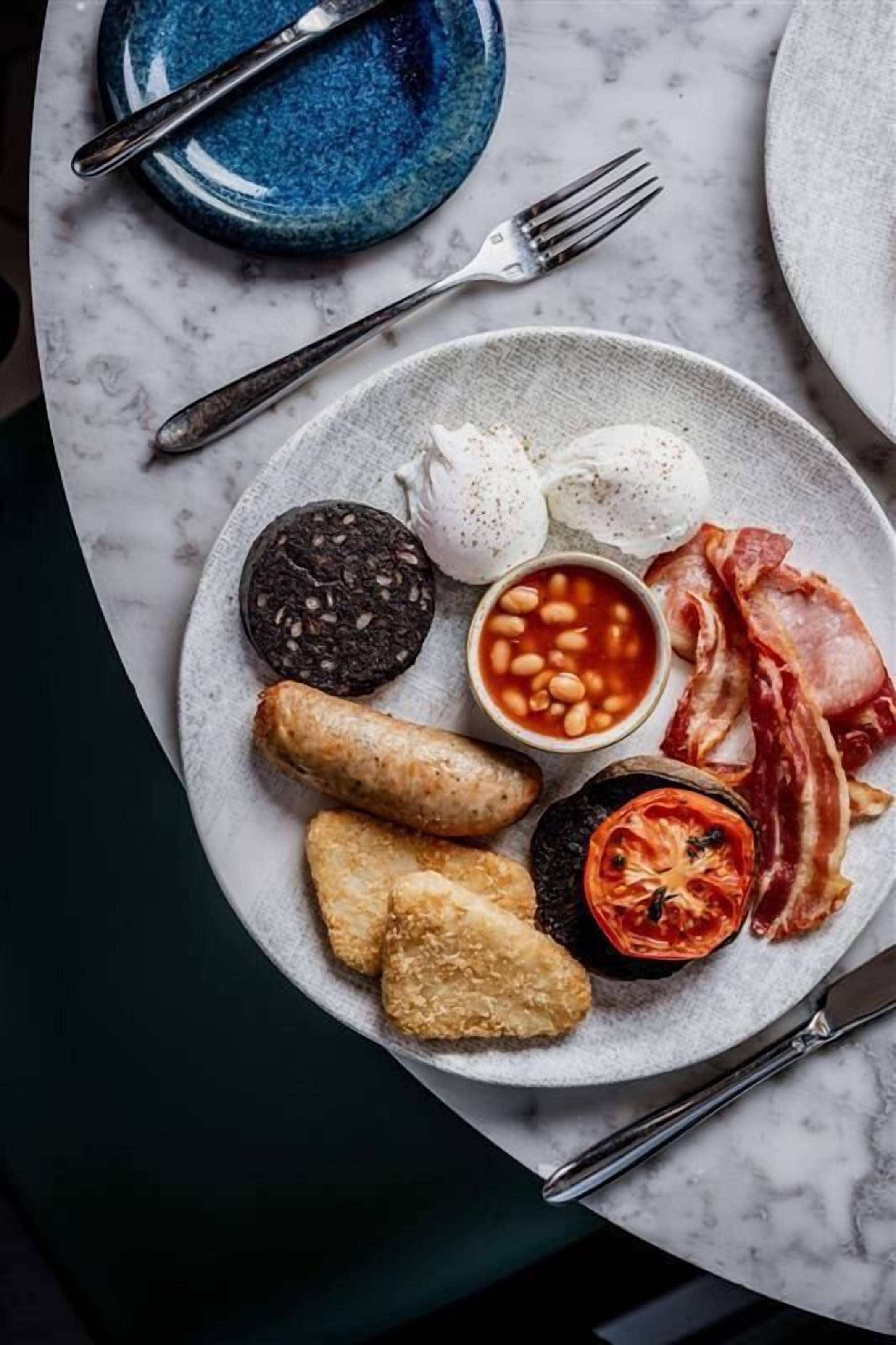 A breakfast plate with eggs, beans, bacon, sausage, and a tomato on a marble table.