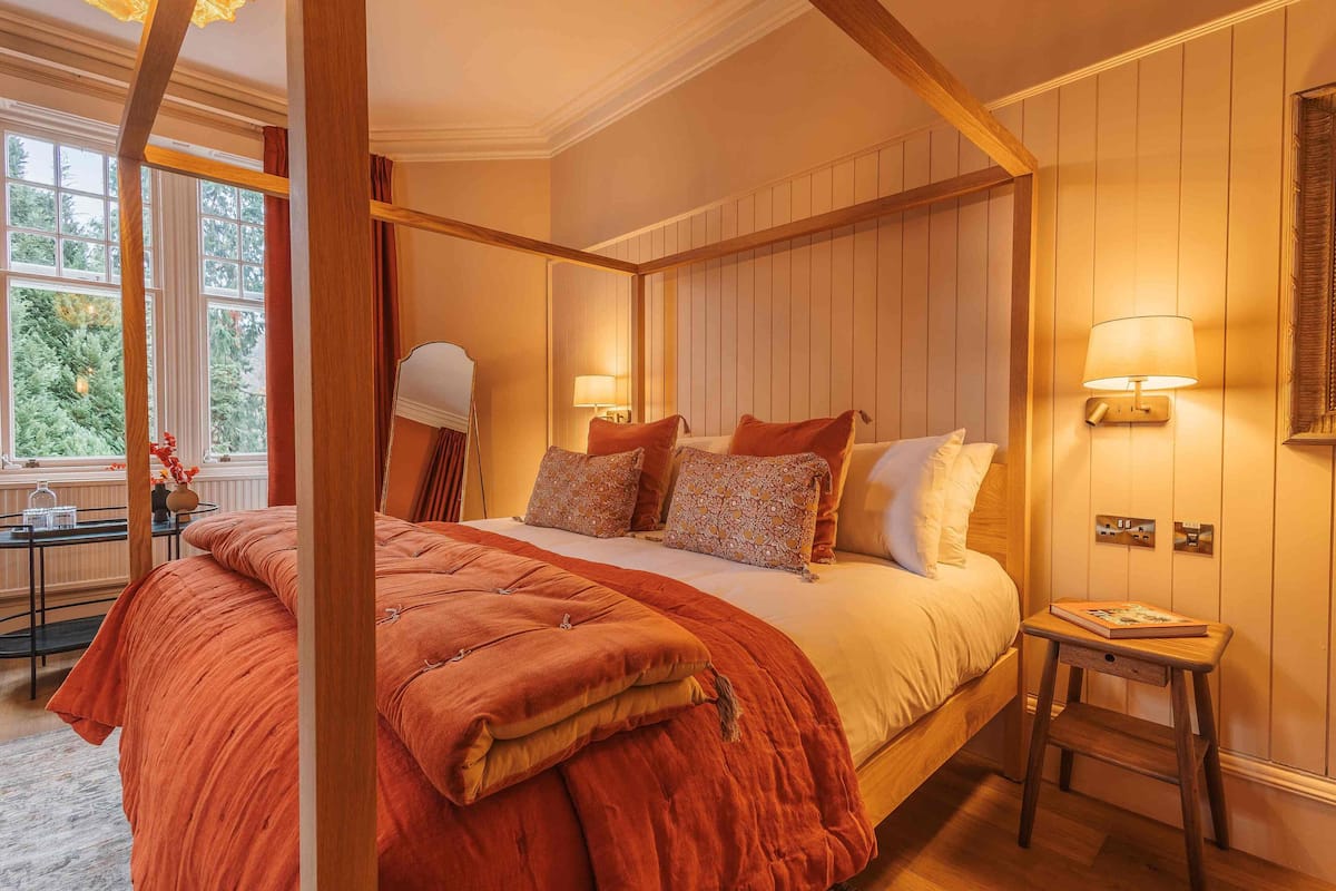 A four-poster bed with a red bedspread, two bedside lamps, and a small wooden nightstand with a book.