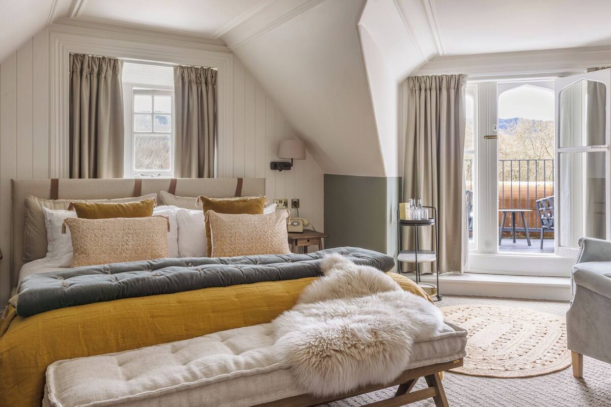 A bedroom with a large bed, a wooden bench, a small table, and a view of the outdoors through a large window.