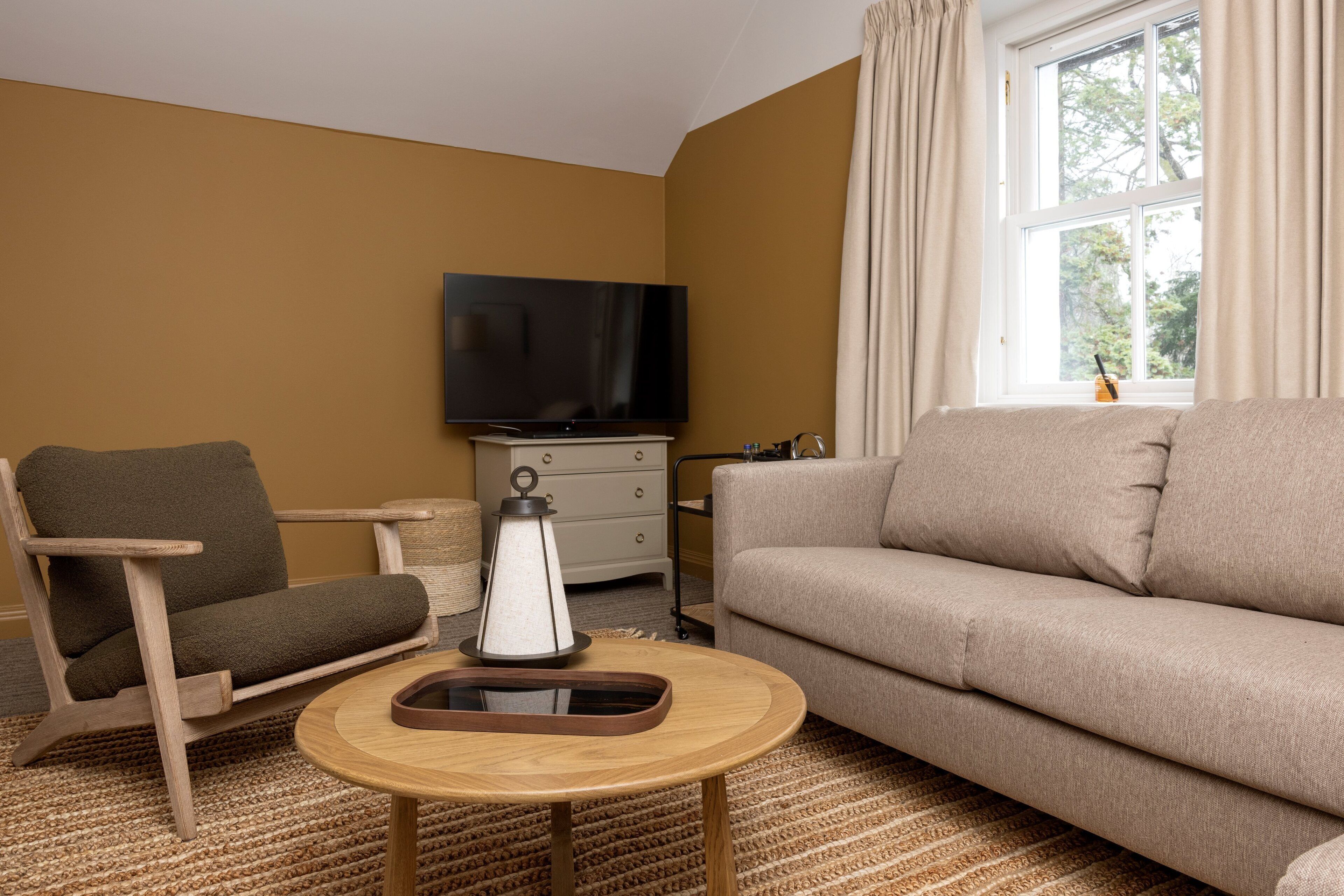 Un salon avec un canapé beige, une chaise en bois, un téléviseur, une petite table avec un plateau, et une fenêtre avec des rideaux.