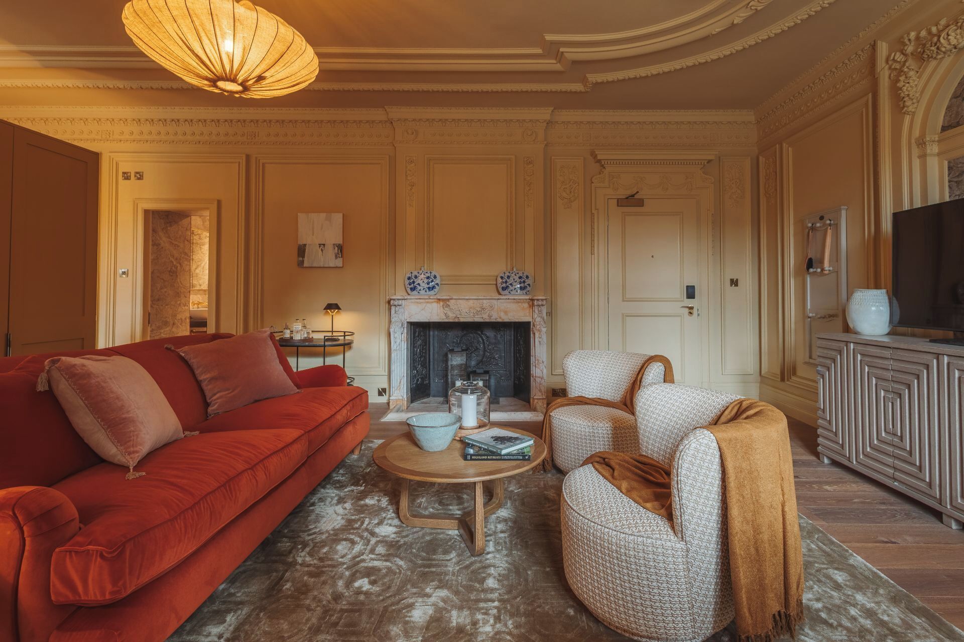 Een woonkamer met een rode bank, twee fauteuils, een salontafel, een open haard en een televisie op een kast.