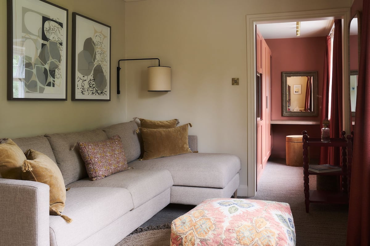 A living room with a sofa, a patterned ottoman, and a hallway leading to a bathroom with a mirror and a toilet.