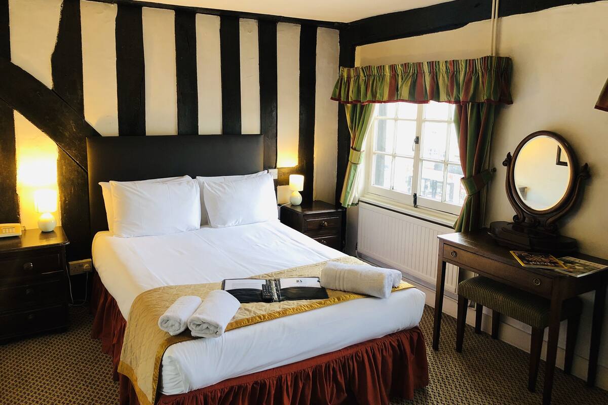 A hotel room with a large bed, a bedside table with a lamp, a mirror, and a window with curtains.