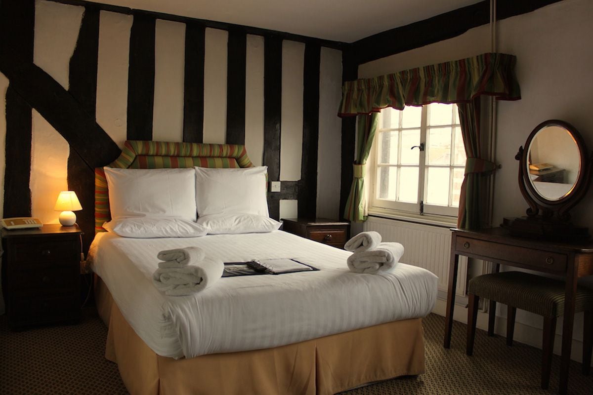 A hotel room with a large bed, a bedside table with a lamp, a dresser, a mirror, and a window with curtains.
