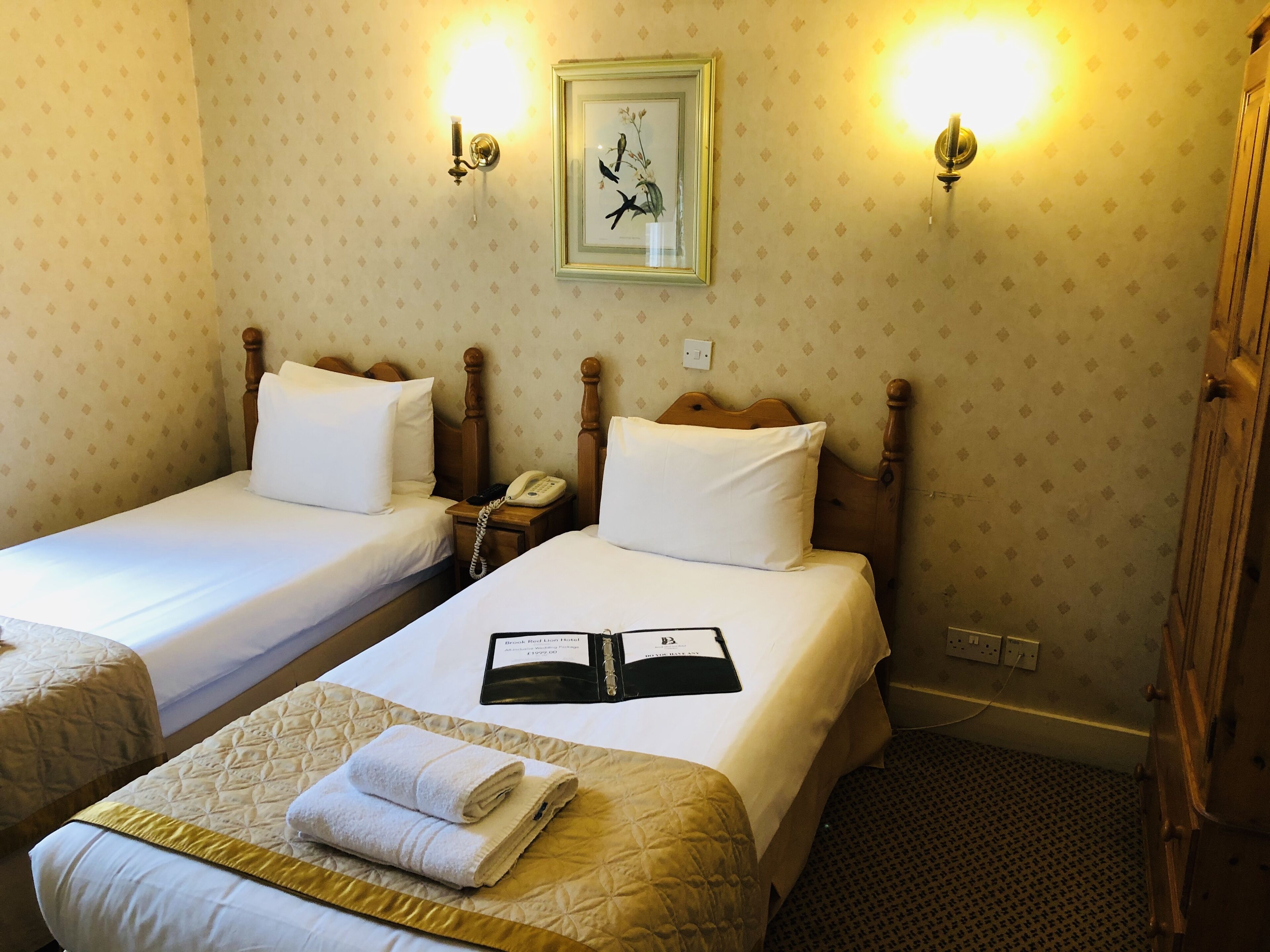 Habitación de hotel con dos camas individuales, una mesita de noche con teléfono, un cuadro en la pared y una cama con un libro y toallas.