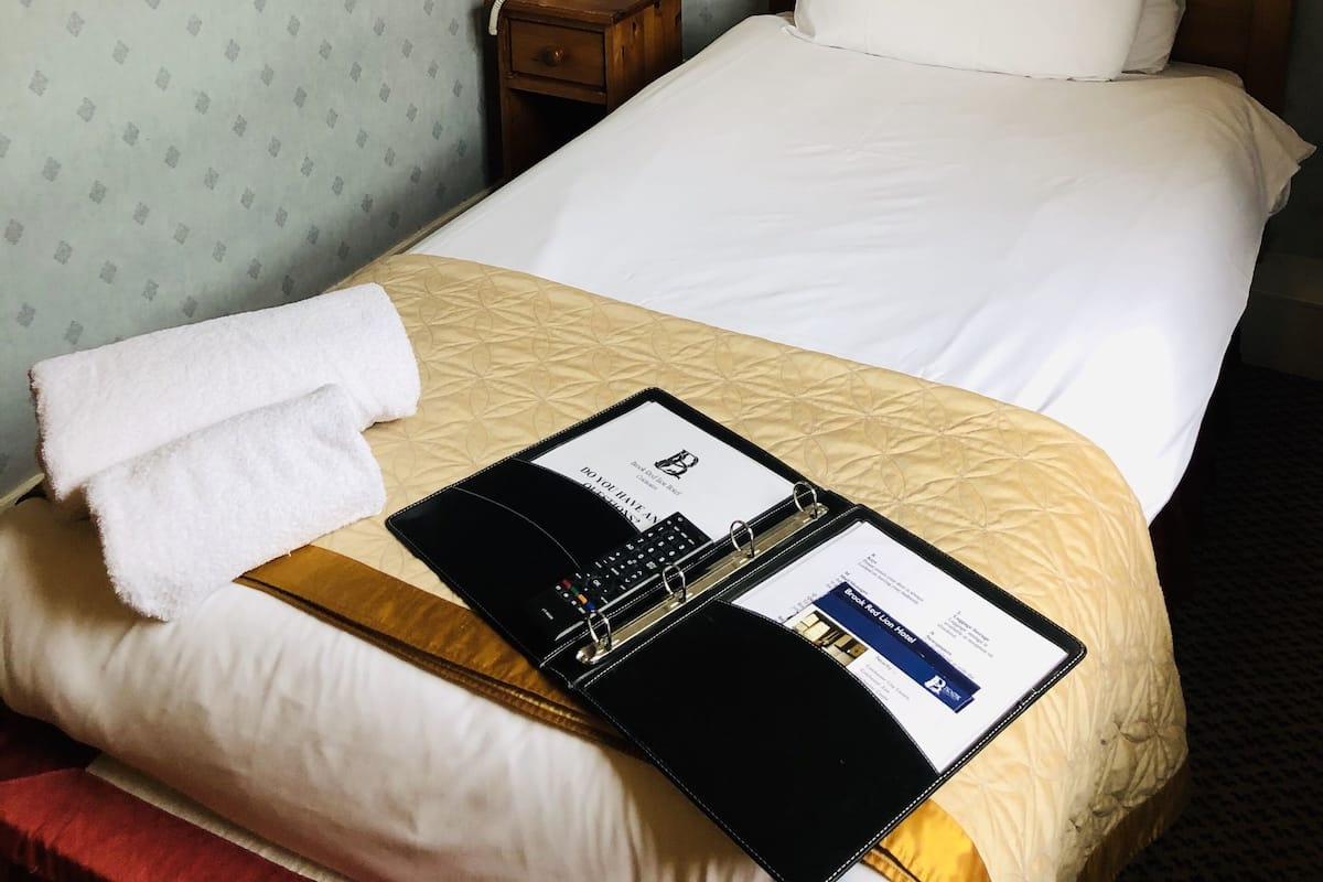 A hotel room with a neatly made bed, a bedside table with a lamp, a framed picture on the wall, and a bulletin board with a map.