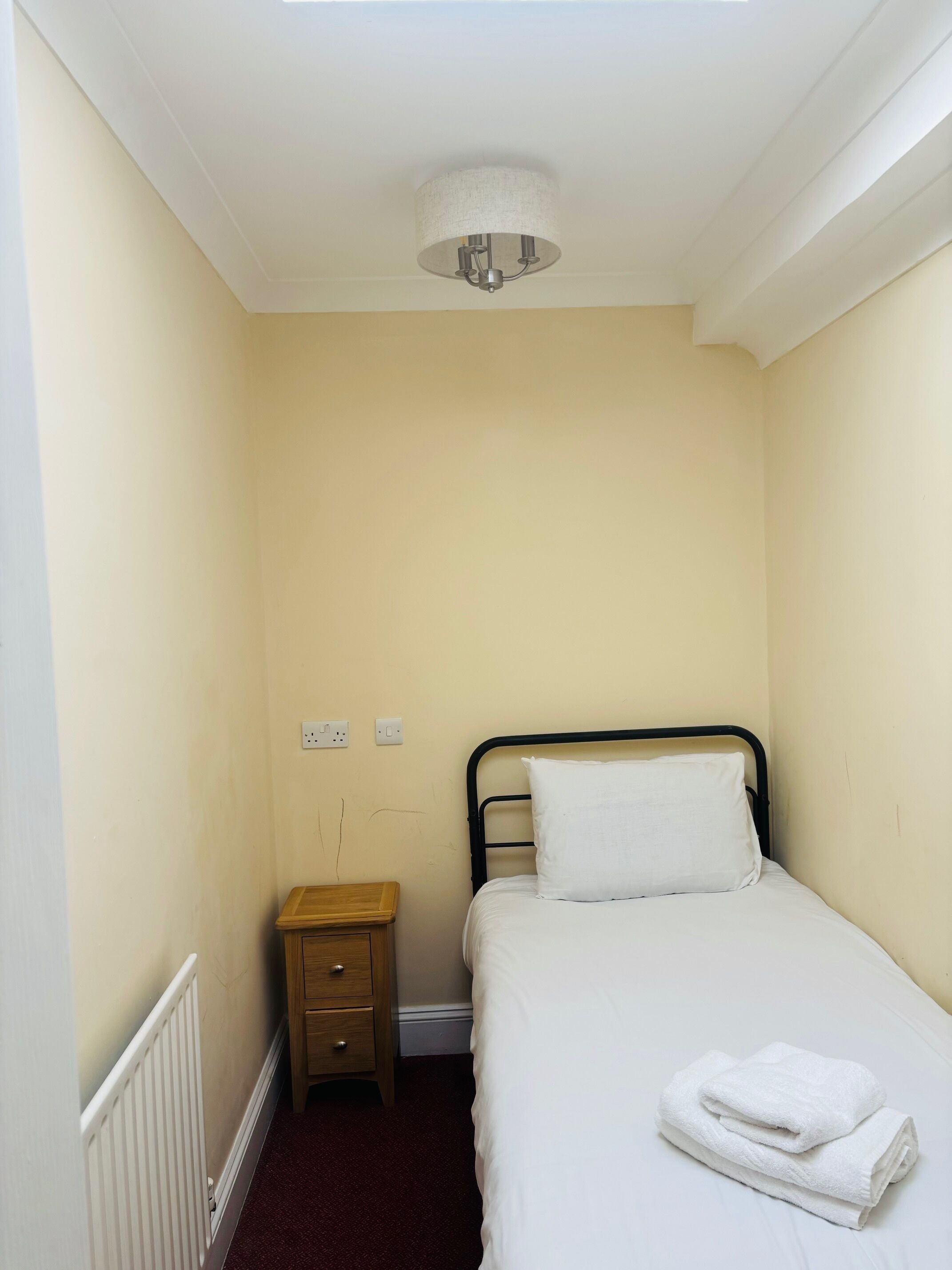 A small, narrow room with a single bed, a nightstand, and a ceiling light fixture.