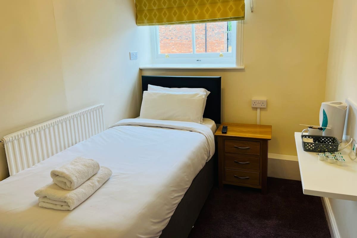 A neatly made bed with white linens and towels, a bedside table with a remote control, a small drawer, and a window with a patterned blind.