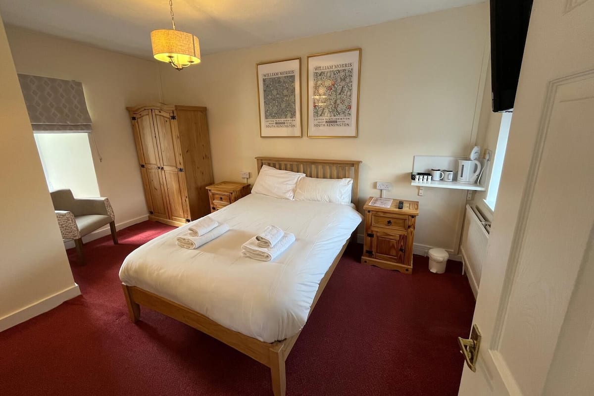 A neatly arranged hotel room with a large bed, a wooden wardrobe, a small bedside table with a lamp, and a window with a view.